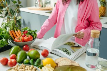A person browsing a cookbook surrounded by fresh vegetables in a bright kitchen setting.